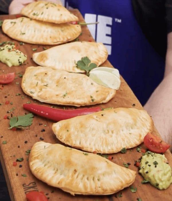 A wooden board holds six golden-brown empanadas garnished with lime, chili pepper, tomato, and a dollop of guacamole, with a person in a blue shirt in the background.