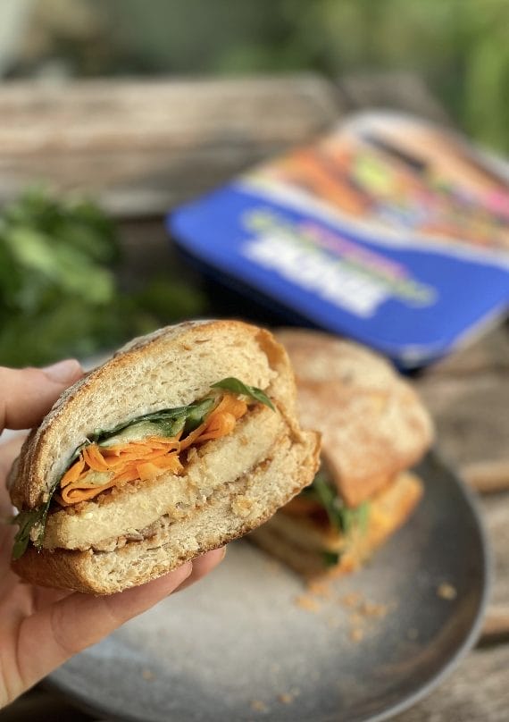 A hand holding a sandwich with carrot, lettuce, and cutlet on a plate, with a blurred notebook in the background.